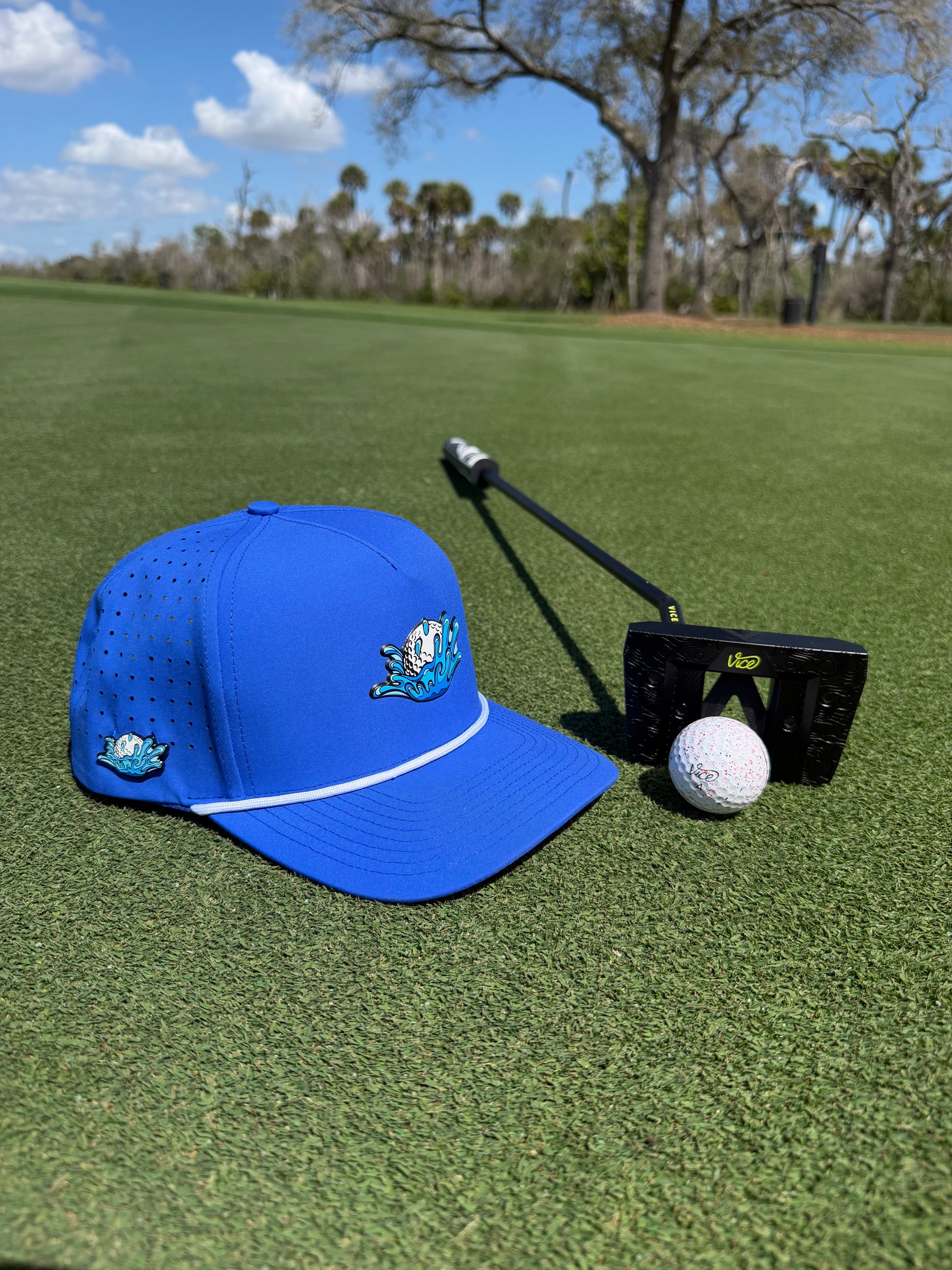 The Water Ball Golf Hat, an adjustable blue snapback cap with built-in magnetic ball marker holder and marker included, sits on green grass beside a golf ball and putter under trees and a clear blue sky.