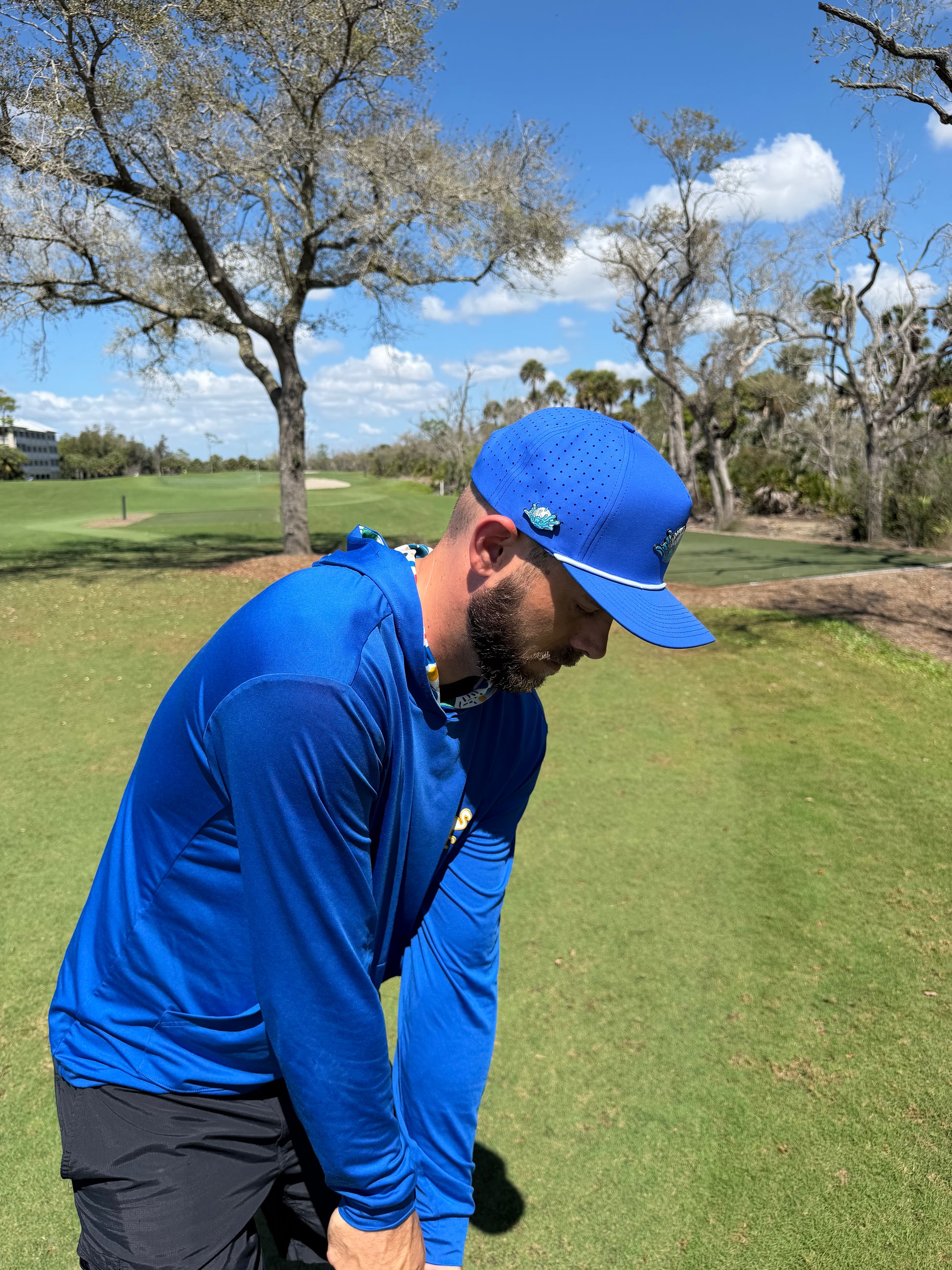 A man wearing the adjustable blue Water Ball Golf Hat, a snapback cap with built-in magnetic ball marker holder and included marker, stands on a green golf course under leafy trees and a blue sky, preparing for his shot.