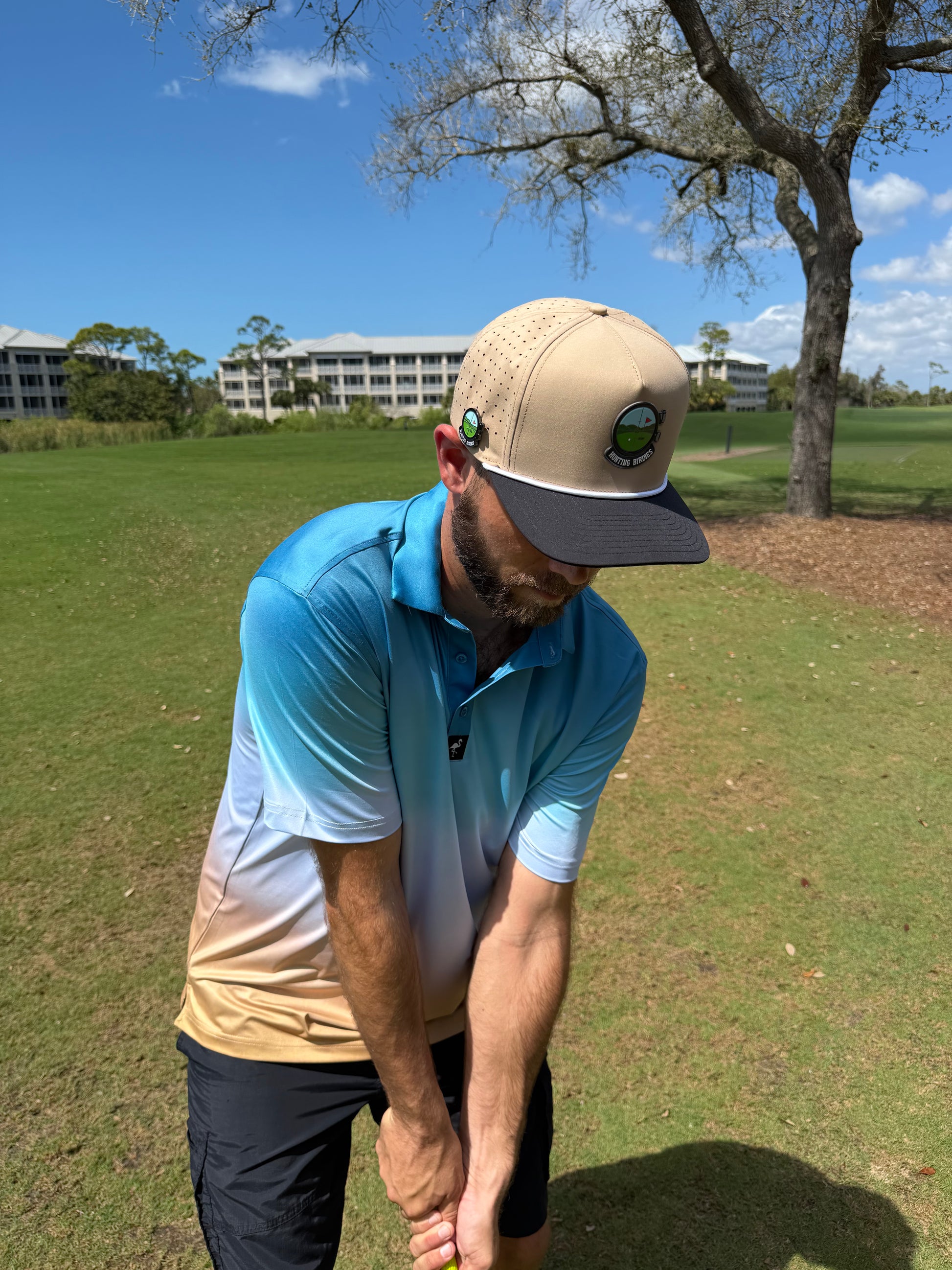 A man in a blue and tan golf shirt and a Hunting Birdies Golf Hat—an adjustable tan & black snapback cap with built-in magnetic ball marker holder—lines up to swing on a sunny, green course with trees and buildings in the background.