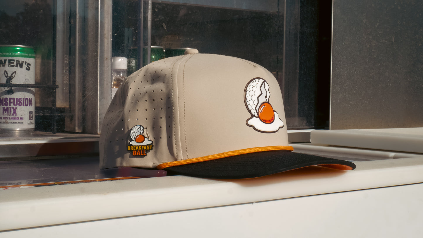 A Mulligan Tan Breakfast Ball Golf Hat with a built-in magnetic ball marker holder and marker, featuring egg and golf ball graphics, text and logo, sits on a white surface by a window.