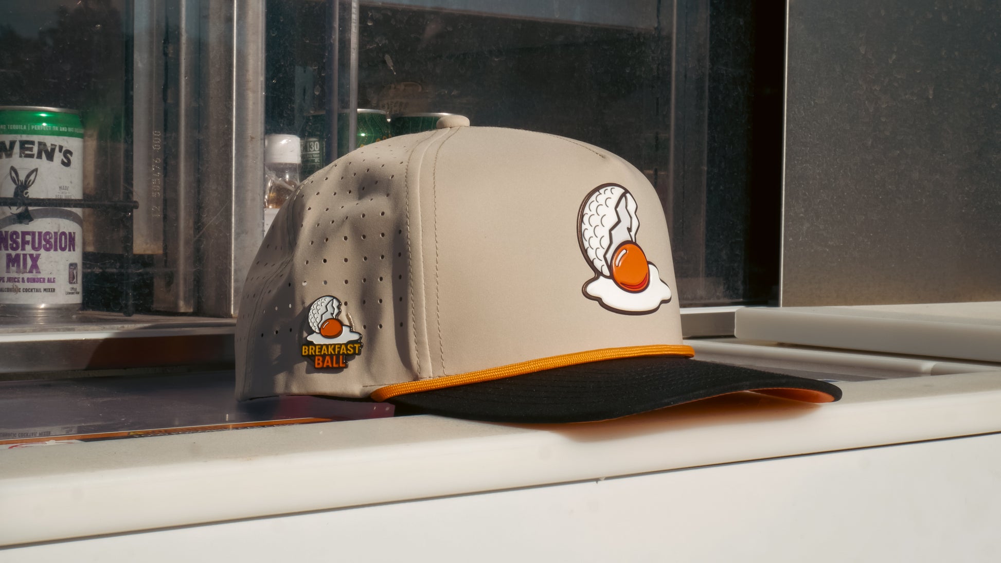 A Mulligan Tan Breakfast Ball Golf Hat with a built-in magnetic ball marker holder and marker, featuring egg and golf ball graphics, text and logo, sits on a white surface by a window.