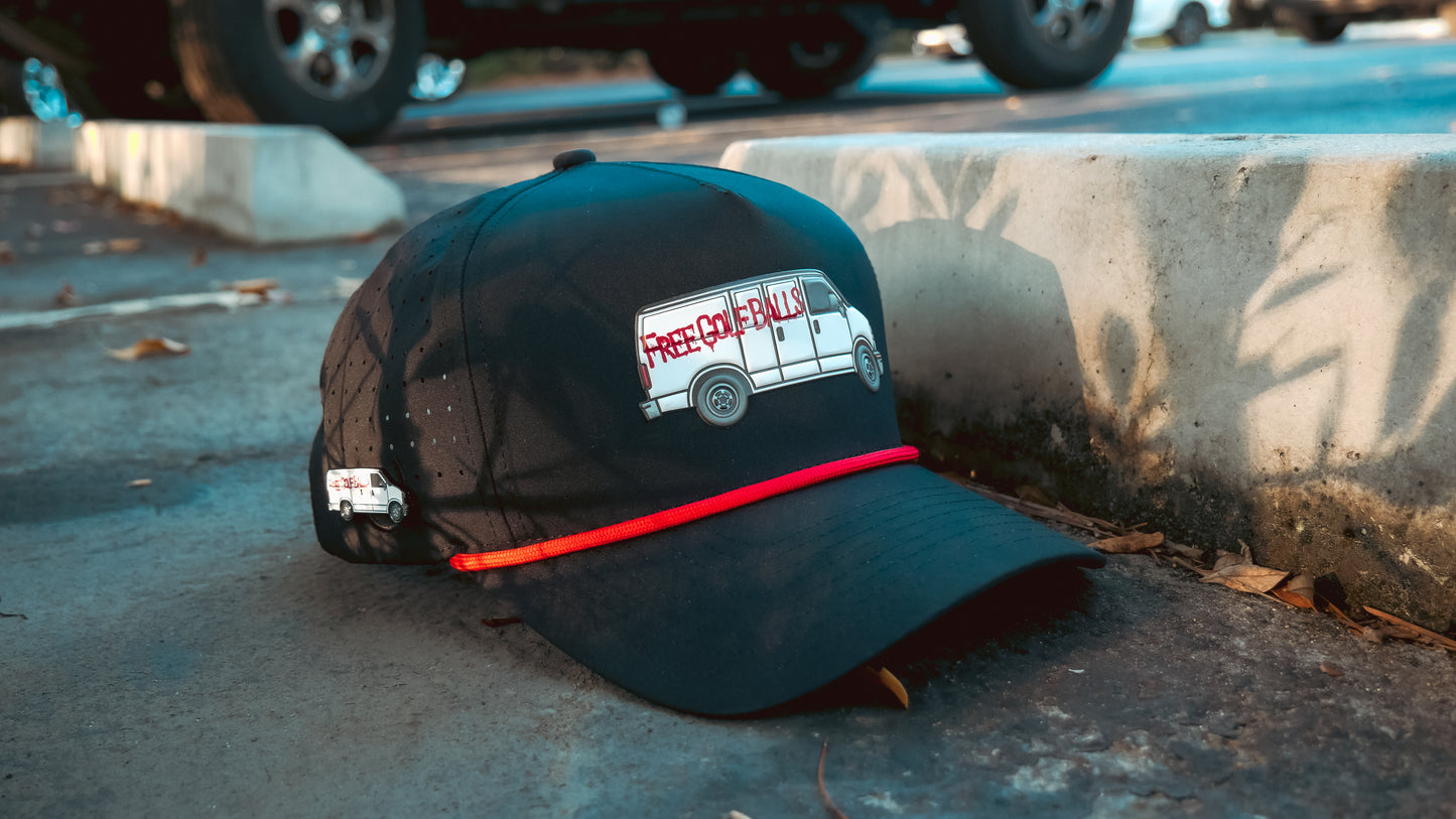 A black Free Golf Balls Van Snapback Cap with red rope detailing sits on the sidewalk. This adjustable golf hat features an embroidered van with "FREE CANDY," a matching pin, and a built-in magnetic ball marker holder with marker included.