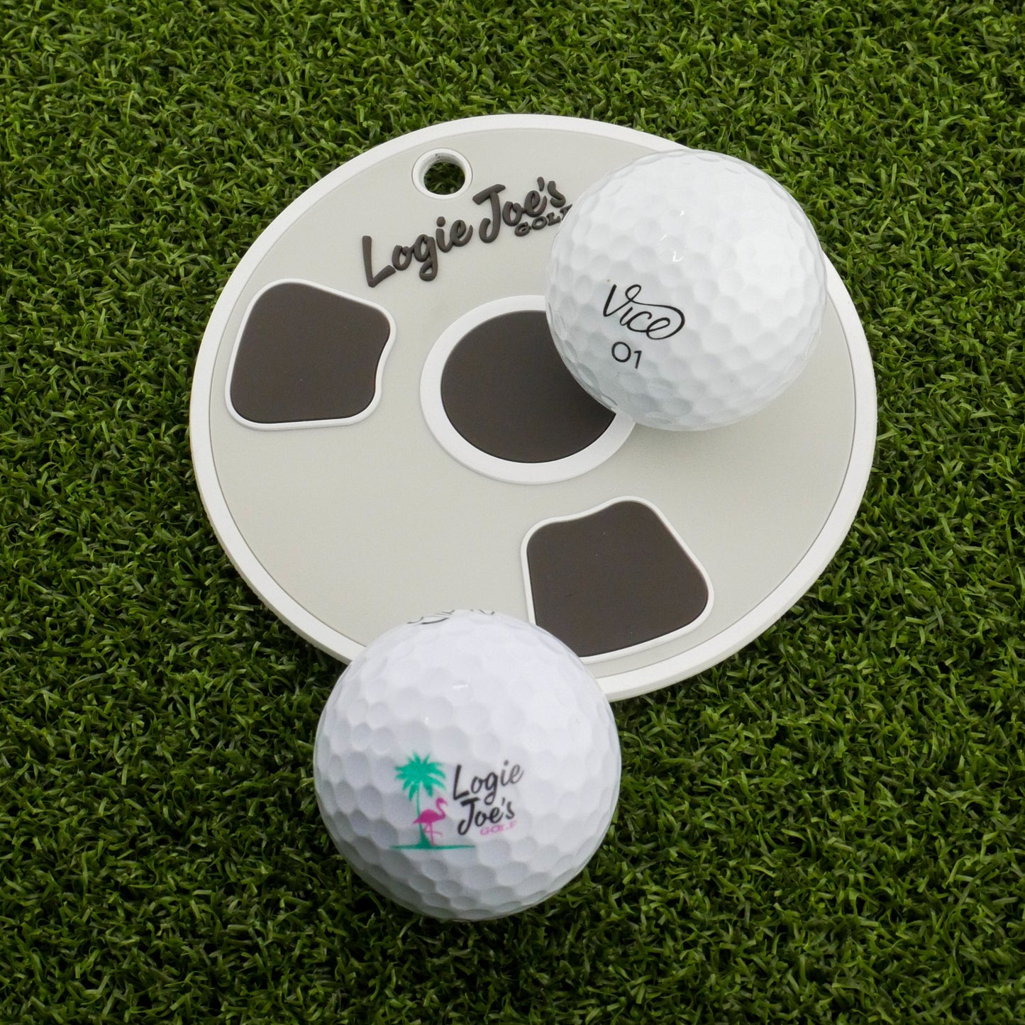 Two white golf balls labeled Logie Joes sit on artificial turf by the Bottom of the Cup Golf Putting Disk, an indoor practice marker and portable training aid featuring the Logie Joes logo—ideal for precision putting practice.