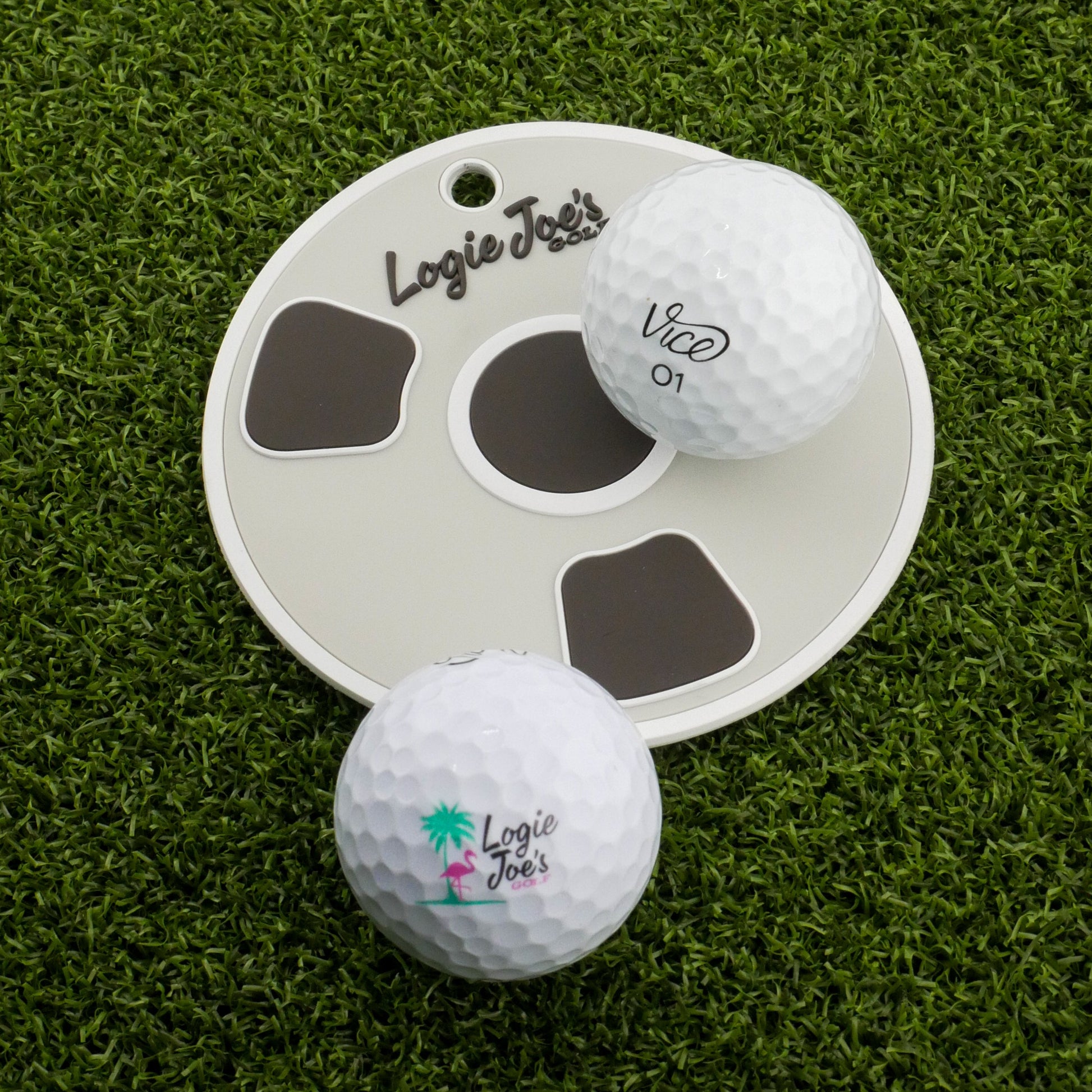 Two white golf balls labeled Logie Joes sit on artificial turf by the Bottom of the Cup Golf Putting Disk, an indoor practice marker and portable training aid featuring the Logie Joes logo—ideal for precision putting practice.