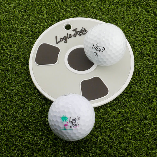 Two white golf balls labeled Logie Joes sit on artificial turf by the Bottom of the Cup Golf Putting Disk, an indoor practice marker and portable training aid featuring the Logie Joes logo—ideal for precision putting practice.