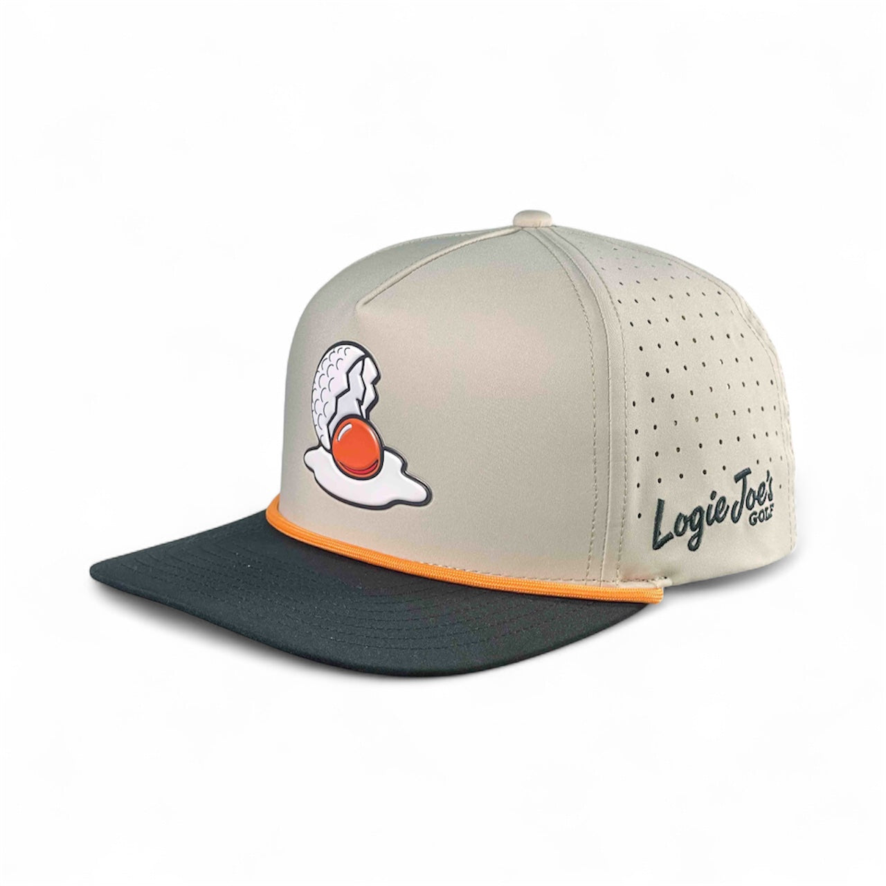 The Breakfast Ball Golf Hat in Mulligan Tan features a cracked egg and yolk design, orange trim, perforated sides, “Logie Joe’s” text, and a built-in magnetic ball marker holder with marker included. Adjustable snapback cap.