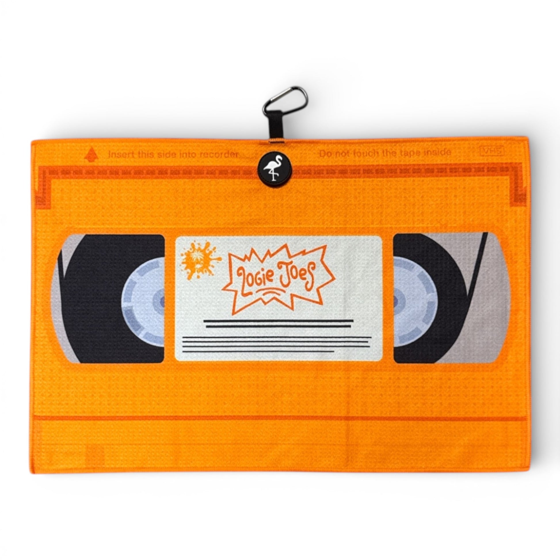 The Nostalgic 90s VHS Golf Towel is an orange, double-sided microfiber towel (24x16 in) with a heavy-duty magnetic carabiner clip, designed to look like a vintage “Logic Joes” VHS tape with illustrated tape reels.