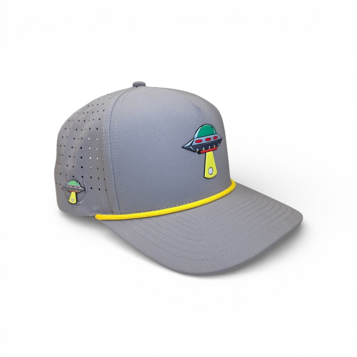 The UFO Golf Hat in Alien Gray features a UFO graphic, yellow trim, ventilation holes, and includes a built-in magnetic ball marker holder with marker. This adjustable snapback cap is the perfect golf essential. Displayed on a white background.