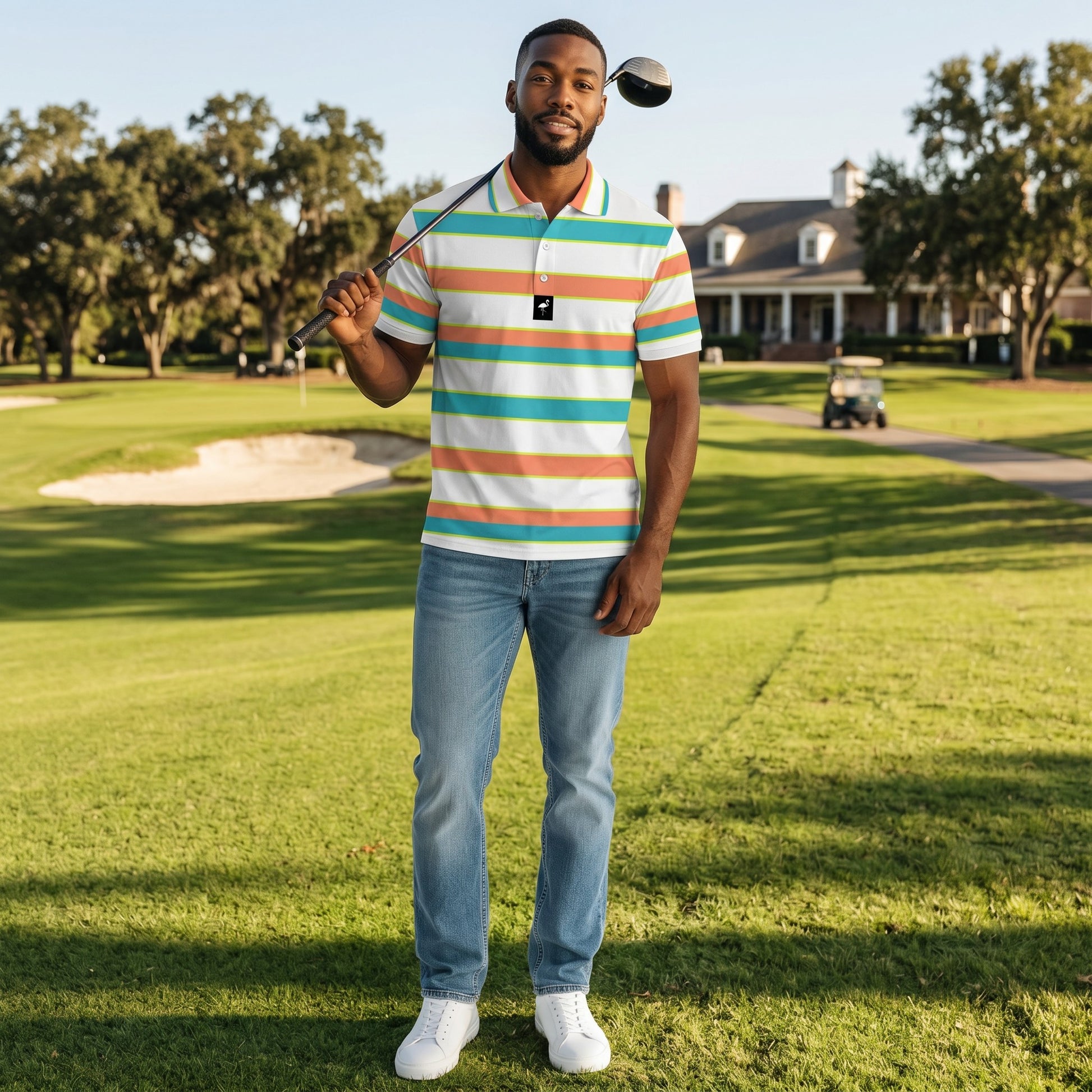 A man stands on a golf course holding a club over his shoulder, wearing the Men's Beach Boardwalk Golf Polo, blue jeans, and white sneakers. A sand trap, trees, and a large house are visible in the background.